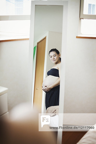 Eine schwangere Frau  die mit den Händen auf dem Bauch vor dem Spiegel steht.