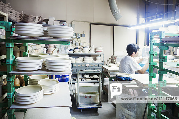 Mann sitzt in einer japanischen Porzellanwerkstatt  umgeben von Regalen mit weißen Porzellantellern  bereit zur Dekoration.