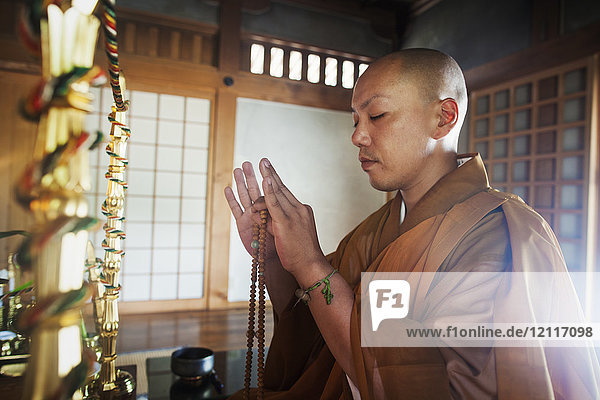 Seitenansicht eines buddhistischen Mönchs mit rasiertem Kopf  der eine goldene Robe trägt  kniend in einem Tempel  Mala haltend  Augen geschlossen.