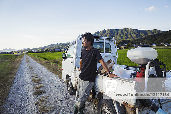 Ein Reisbauer steht an seinem Lastwagen und schaut über die flache Landschaft  die Reisfelder und ein Dorf in der Ferne.
