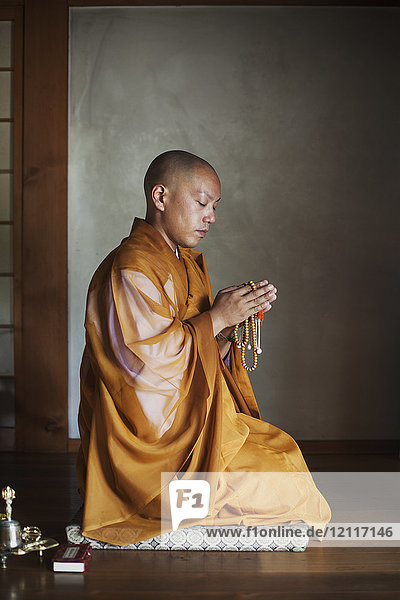 Seitenansicht eines buddhistischen Mönchs mit rasiertem Kopf  der in einem goldenen Gewand in einem Tempel kniend  Mala haltend und betend in einem Tempel sitzt.