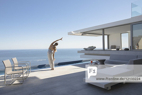 Frau übt Yoga Seite dehnen auf moderne  luxuriöse Haus Schaufenster Außenterrasse mit sonnigen Blick auf das Meer