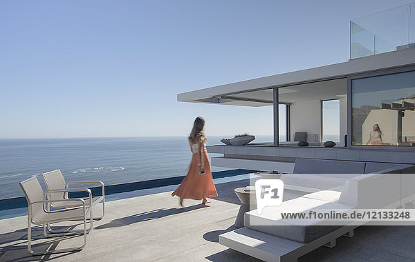 Frau im Kleid  die auf einer sonnigen  modernen  luxuriösen Wohnung spazieren geht  die eine Außenterrasse mit Meerblick hat