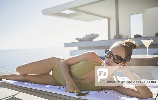 Porträt selbstbewusste Frau im Badeanzug beim Sonnenbaden auf sonniger Luxus-Terrasse  Musik hören mit Smartphone