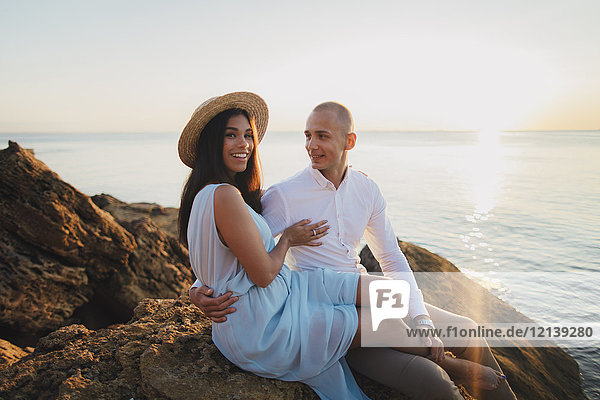 Caucasian couple sitting on rocks on beach at sunset