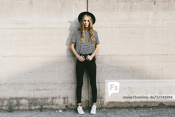 Fashionable young woman wearing hat leaning against concrete wall