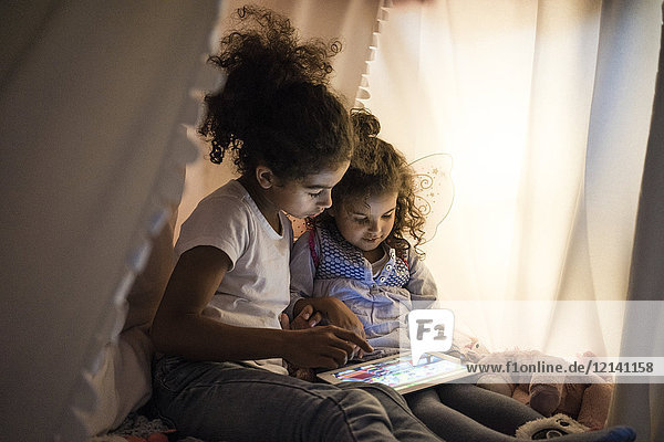 Zwei Schwestern sitzen im dunklen Kinderzimmer und schauen auf das digitale Tablett.