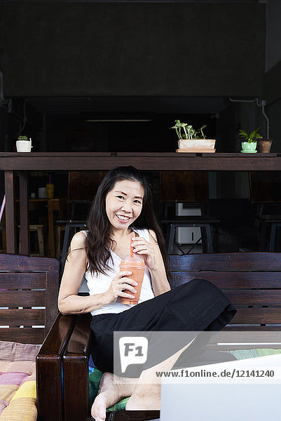 Porträt einer lächelnden Frau  die auf einer Terrassenbank sitzt und einen Smoothie trinkt.