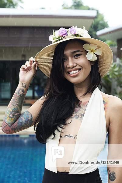 Porträt einer lächelnden Frau mit Blume im Haar  die einen Strohhut am Swimmingpool trägt.
