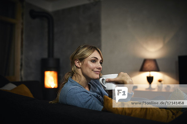 Porträt einer glücklichen Frau mit einer Tasse Tee  die abends zu Hause auf der Couch liegt.