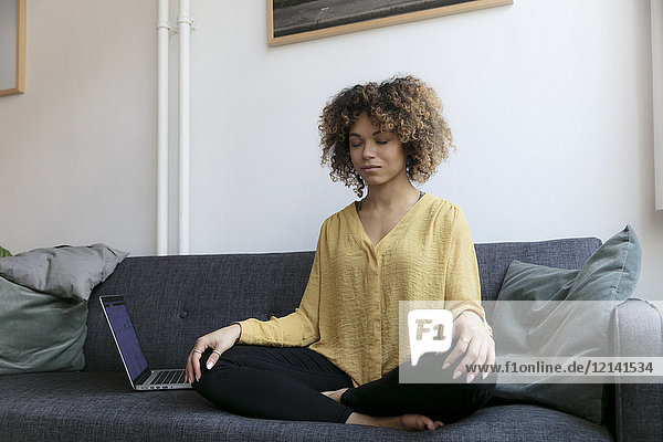 Young woman sitting on couch at home next to laptop meditating