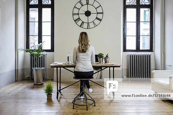 Rear view of woman sitting at desk at home under large wall clock