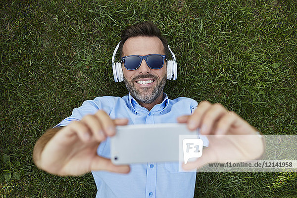 Porträt eines lachenden Mannes auf einer Wiese liegend  Selfie mit Smartphone  Draufsicht