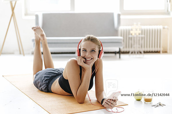 Smiling young woman in sportswear lying on gym mat listening to music
