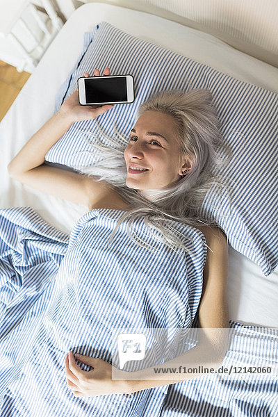 Smiling young woman lying in bed holding cell phone