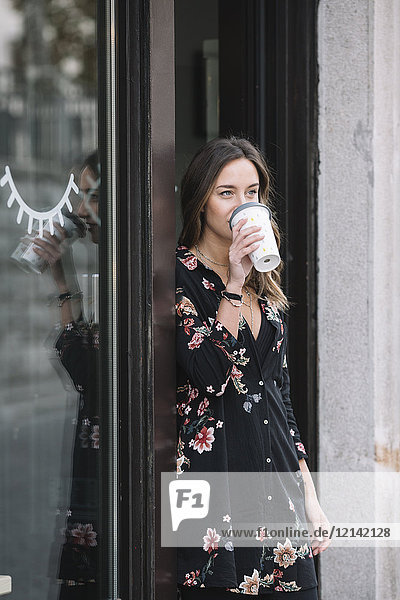 Modische Frau lehnt sich an die Eingangstür ihres Ladens und trinkt Kaffee zum Mitnehmen.