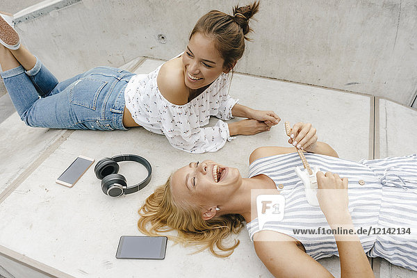 Zwei glückliche junge Frauen  die auf einer Rampe in einem Skatepark liegen und Musik machen.