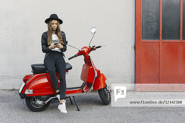 Modische junge Frau sitzend auf rotem Motorroller mit Handy