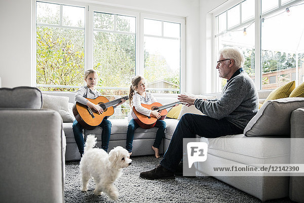 Zwei Mädchen auf dem Sofa spielen Gitarre mit Großvater und Hund