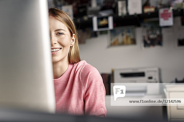 Porträt der lächelnden jungen Frau hinter dem Computerbildschirm am Schreibtisch zu Hause