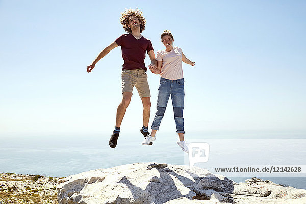 South Africa  Cape Town  happy young couple jumping on top of a mountain at the coast