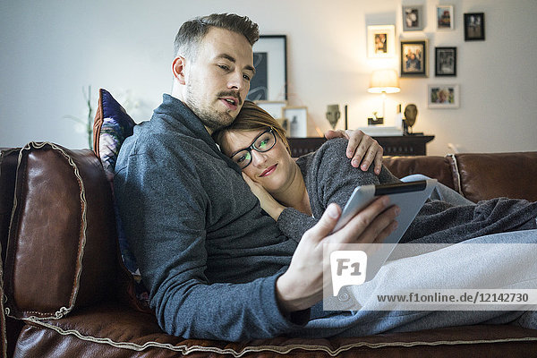 Lächelndes Paar liegt auf der Couch zu Hause und teilt sich das Tablett.