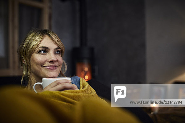 Porträt einer lächelnden Frau mit einer Tasse Kaffee,  die abends zu Hause auf der Couch liegt.