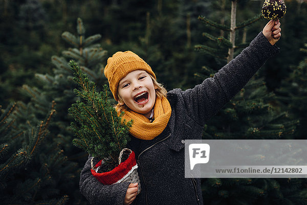 Fröhlicher Junge bereitet sich auf Weihnachten vor  hält Topfbaum  isst Schokolade getauchten Apfel