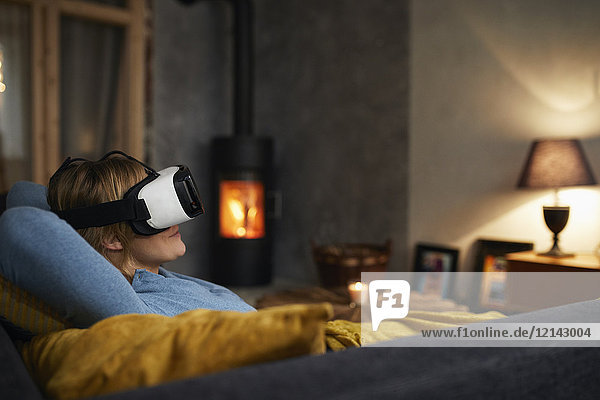Frau  die abends zu Hause auf der Couch liegt und eine Virtual Reality Brille benutzt.
