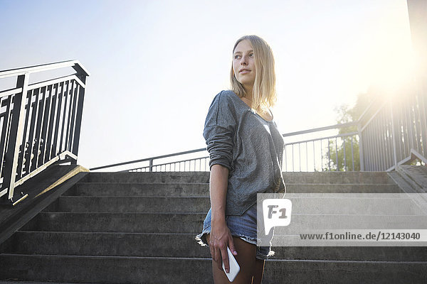 Blonde junge Frau mit Smartphone im Gegenlicht vor der Treppe stehend
