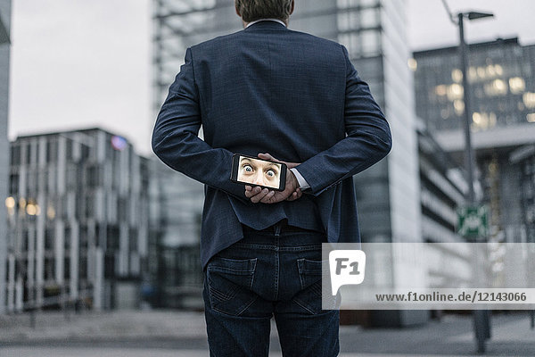 Businessman holding cell phone with image of eyes behind his back