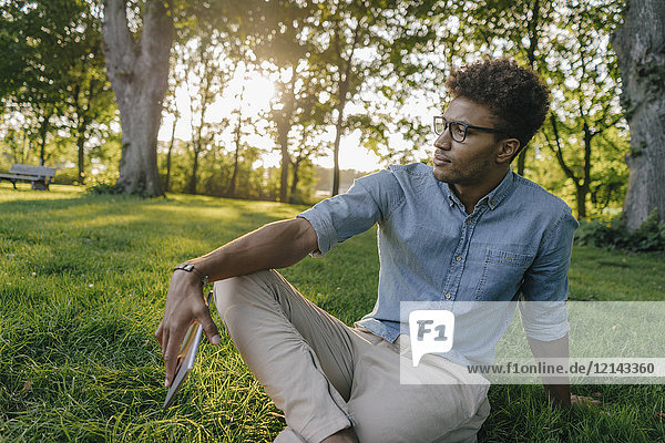 Junger Mann sitzt mit mobilem Gerät im Park und schaut sich um