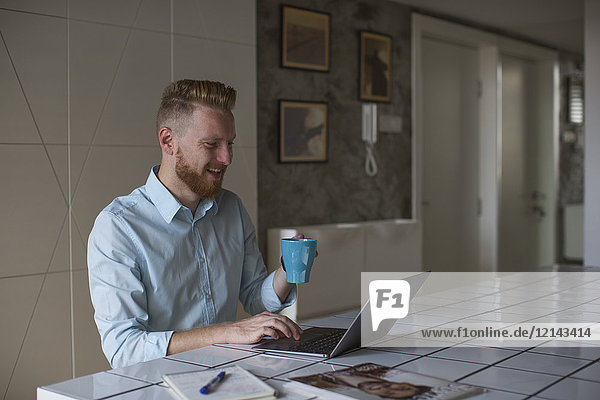Lachender Geschäftsmann mit Kaffeetasse bei der Arbeit an seinem Laptop zu Hause
