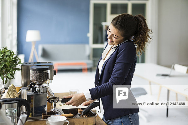 Lächelnde Geschäftsfrau am Telefon beim Zubereiten von Espresso mit Espressomaschine im Loft