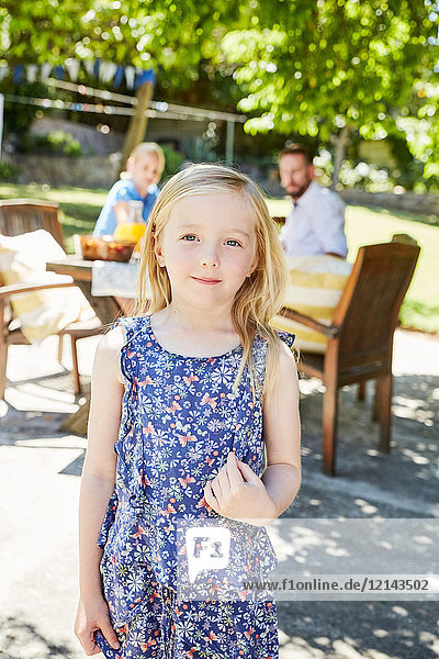 Porträt eines lächelnden Mädchens mit Eltern im Hintergrund am Gartentisch