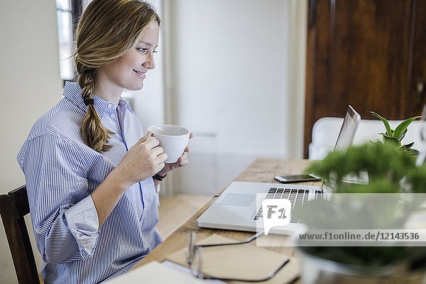 Smiling woman sitting at desk with cup of coffee and laptop