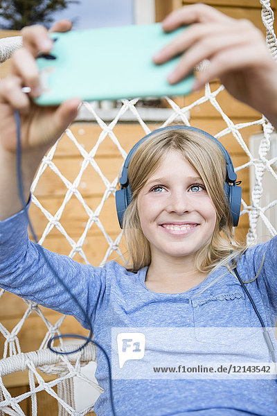Porträt eines lachenden Mädchens mit Kopfhörern  das sich selbst mit einem Smartphone in einem Hängesessel nimmt.