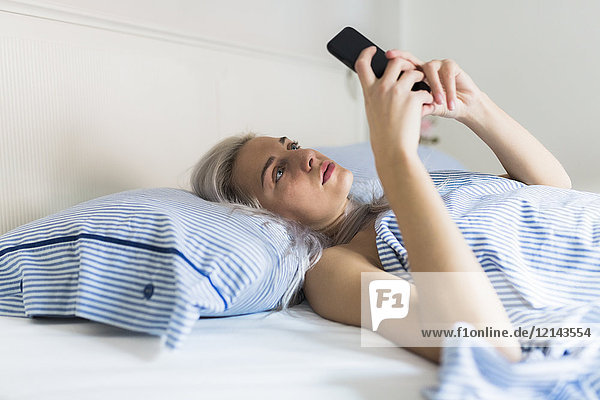 Seriöse junge Frau liegt im Bett und überprüft ihr Handy.