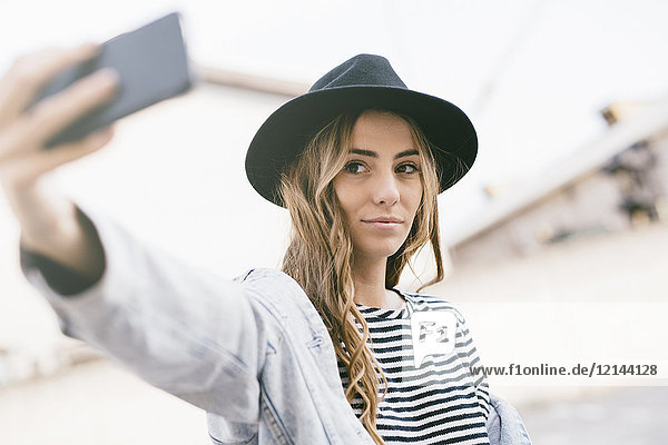 Porträt einer modischen jungen Frau  die einen Hut trägt und sich selbst mit dem Smartphone trägt.