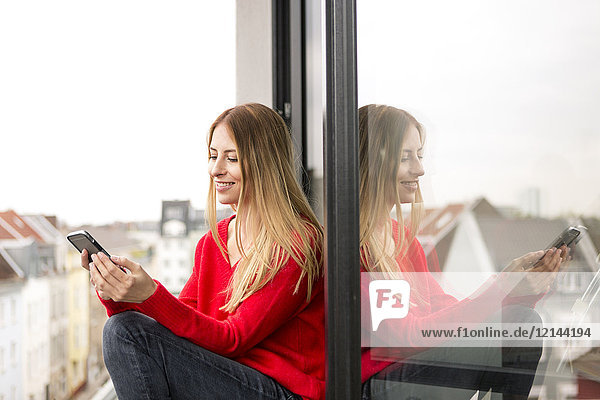 Lächelnde junge Frau am Fenster in der Stadtwohnung mit Blick auf das Handy