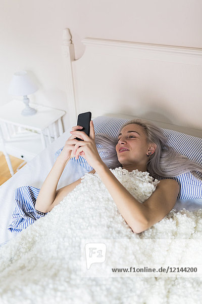 Smiling young woman lying in bed checking cell phone