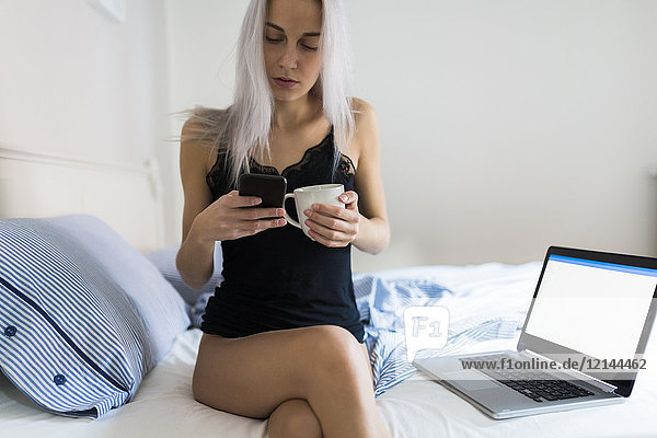 Young woman in bed with cell phone  cup of coffee and laptop