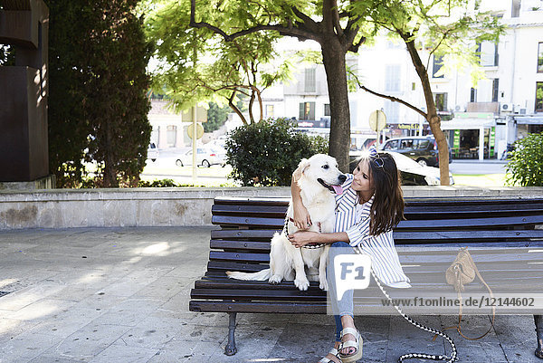 Happy young woman sitting with her dog on bench in the city