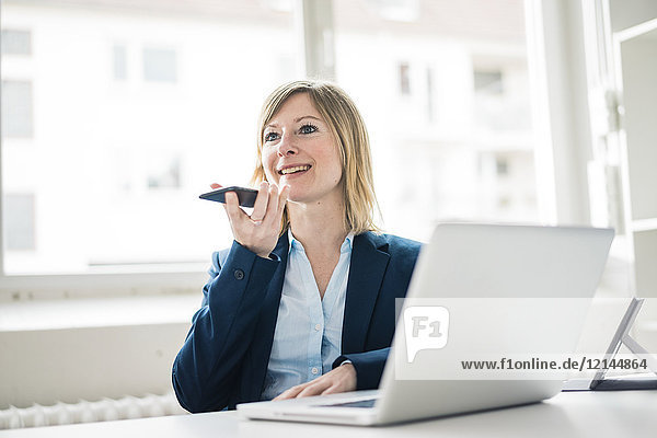 Lächelnde Geschäftsfrau im Büro mit Handy und Laptop