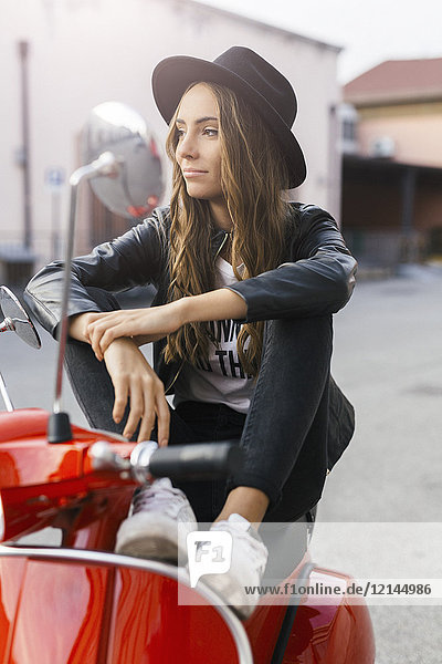 Porträt der modischen jungen Frau auf rotem Motorroller sitzend
