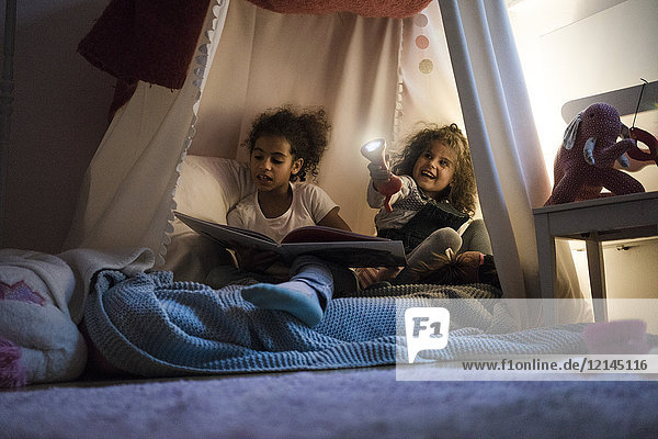 Zwei Schwestern sitzen im dunklen Kinderzimmer  spielen mit Fackel  lesen Buch