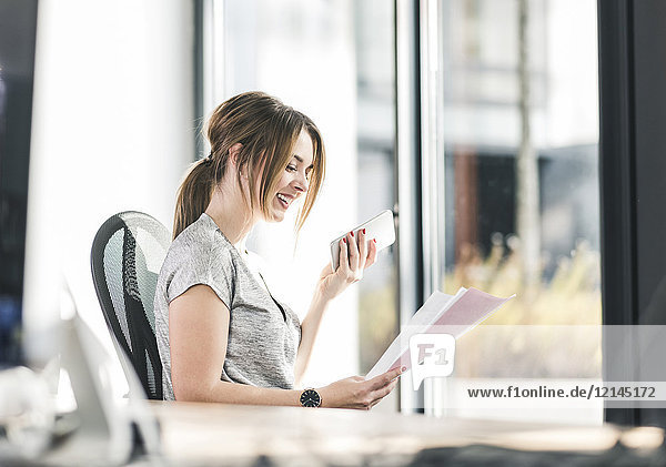 Smiling businesswoman with cell phone and documents at desk in office