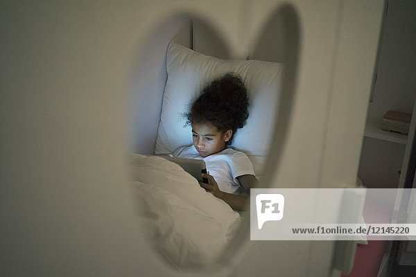 Kleines Mädchen sitzt im dunklen Kinderzimmer und schaut auf ein digitales Tablett.