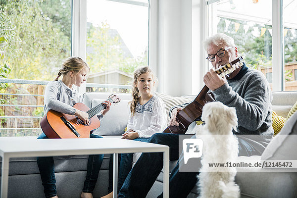 Two girls and grandfather on sofa playing guitar with dog watching