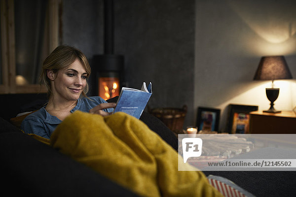 Porträt einer lächelnden Frau beim Lesen eines Buches auf der Couch zu Hause am Abend